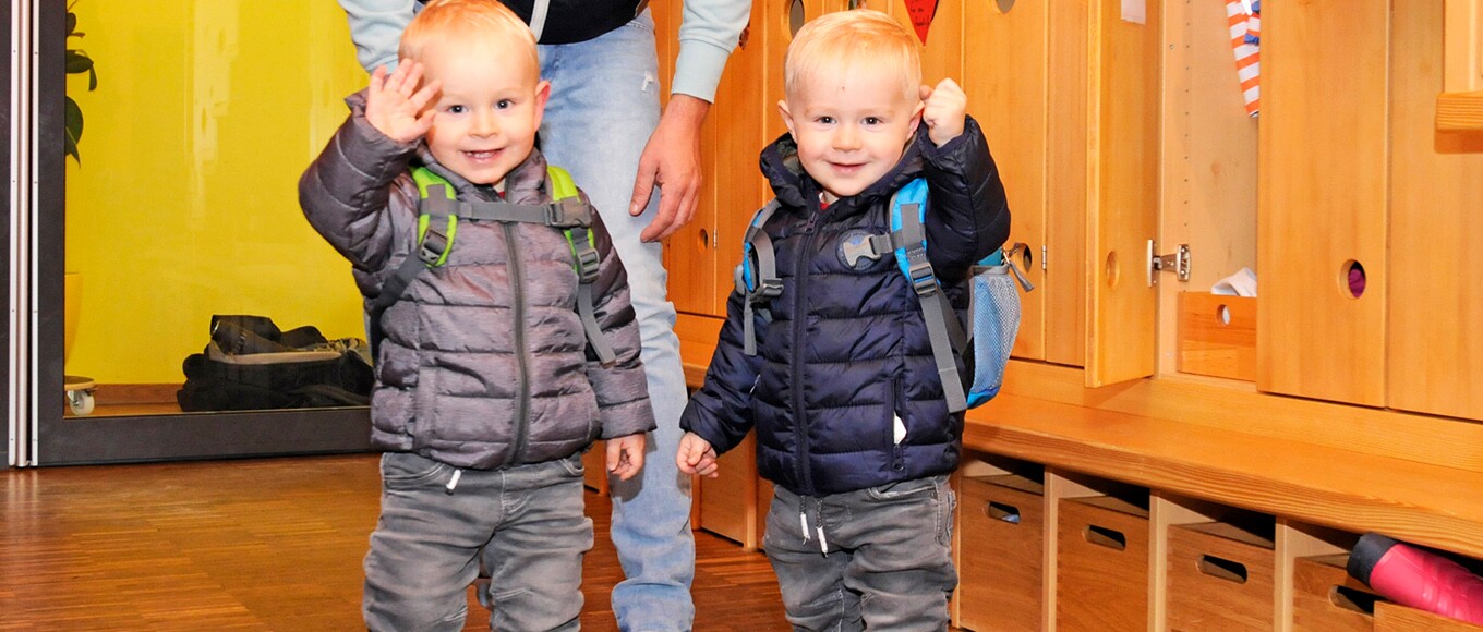 Zwei kleine Buben mit Rucksäcken winken in die Kamera