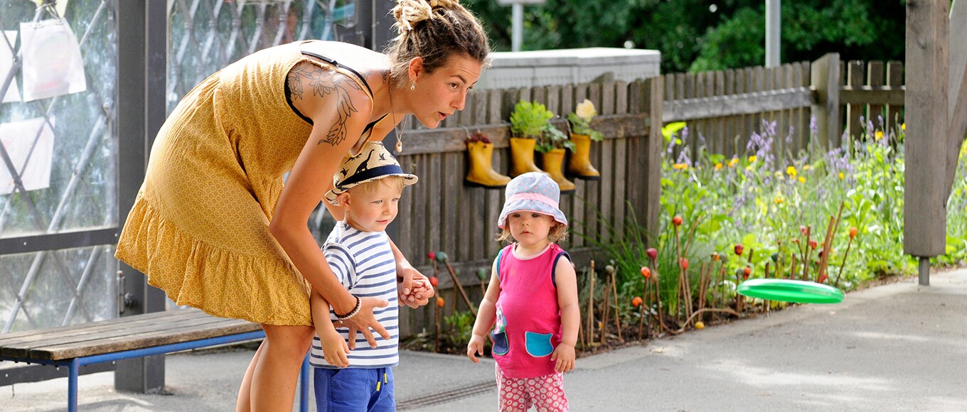 Betreuerin mit Dreadlocks in einem gelben Sommerkleid mit zwei Kleinkindern im blühenden Garten